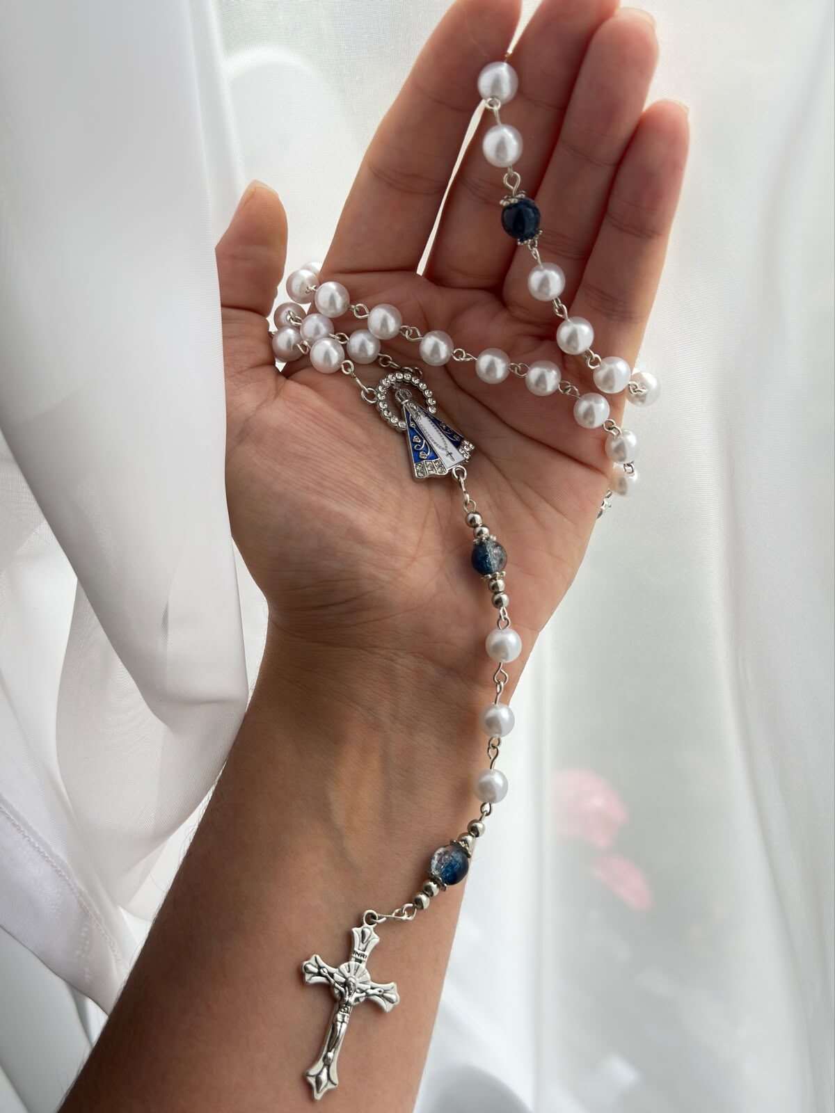Small round white beads on pale-toned wire links lying open across a palm, with deep blue beads at the larger positions and a blue-and-white enamelled standing figure at the centre