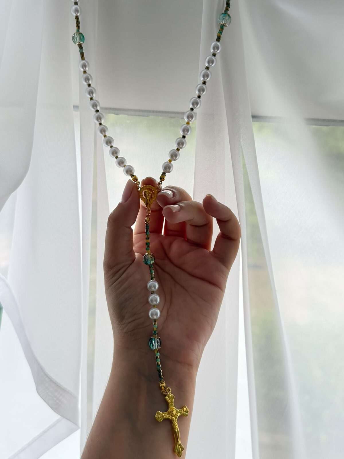 A long strand of small round white beads hanging at full length from raised fingers against a backlit curtain, green-blue notes recurring along it and a warm-toned figured cross at the foot