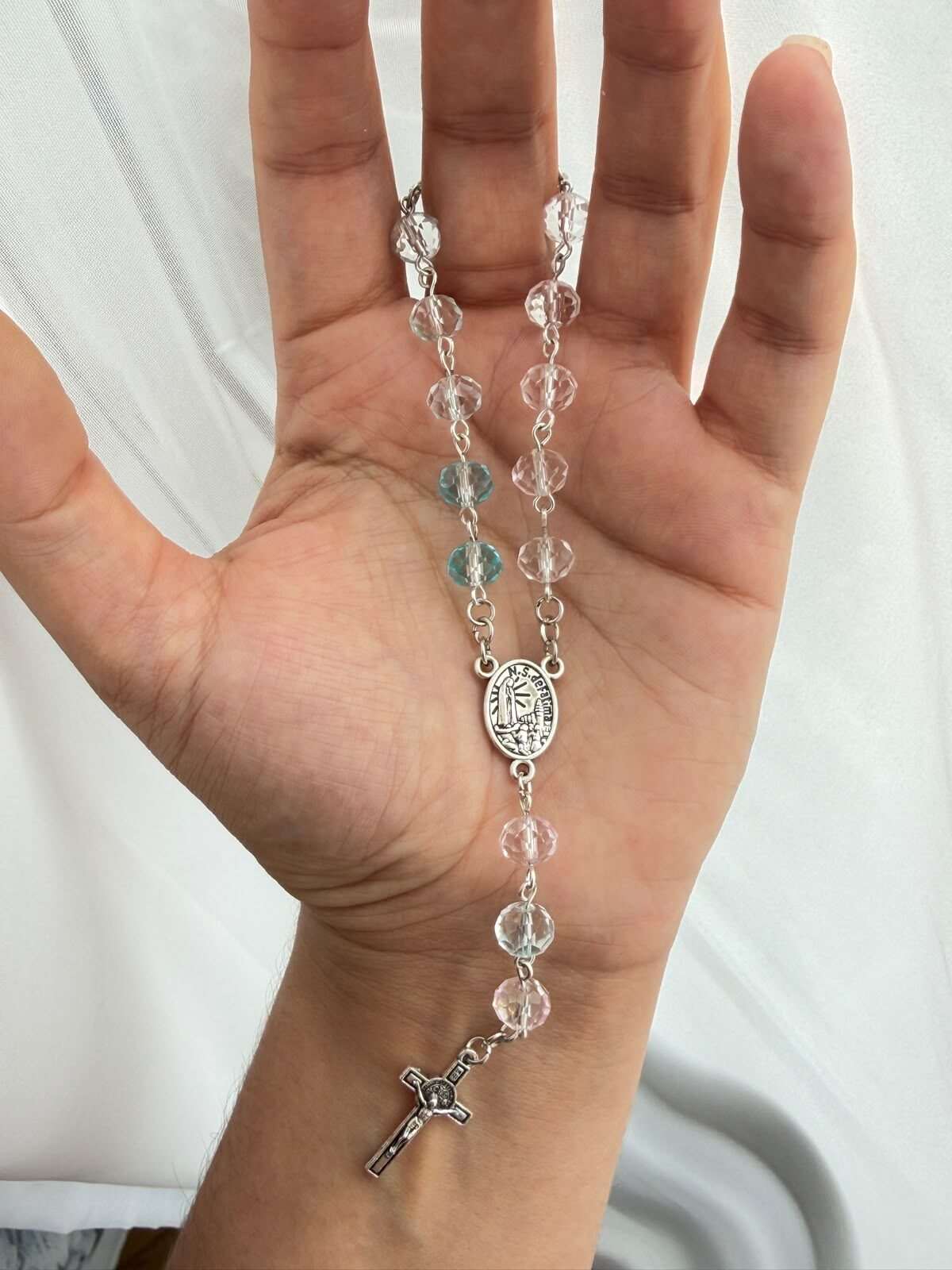 Colourless faceted beads on fine pale-toned wire links lying open across a palm, two pale aqua beads low in the run, an oval centre medal and a plain cross at the foot