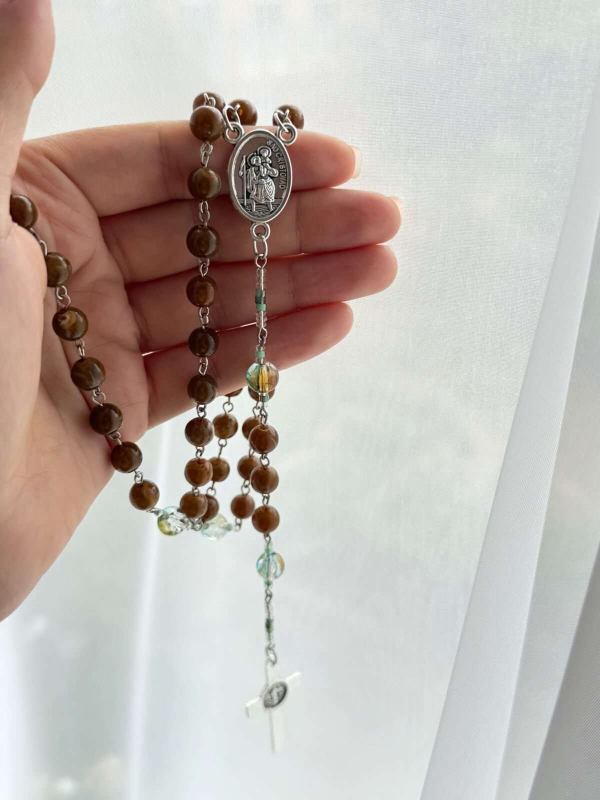 Brown marbled beads on fine pale-toned wire links resting across an open palm, small green and amber notes at the turns, an oval medal showing a figure carrying a child, and a plain white cross below