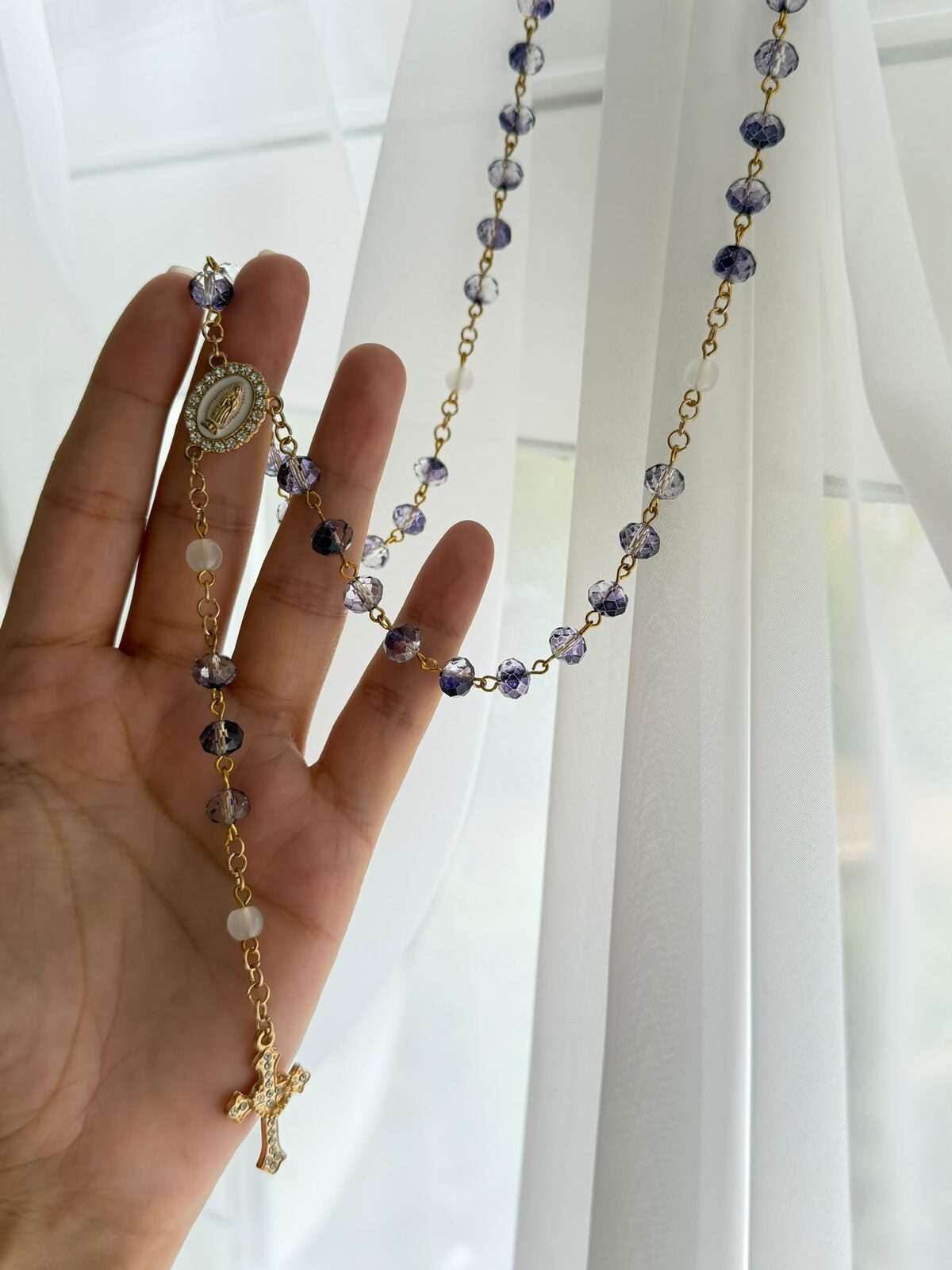 Deep violet faceted beads on warm-toned wire links falling across an open palm, a milky white note at each turn, a small oval centre and a slender cross set with clear points