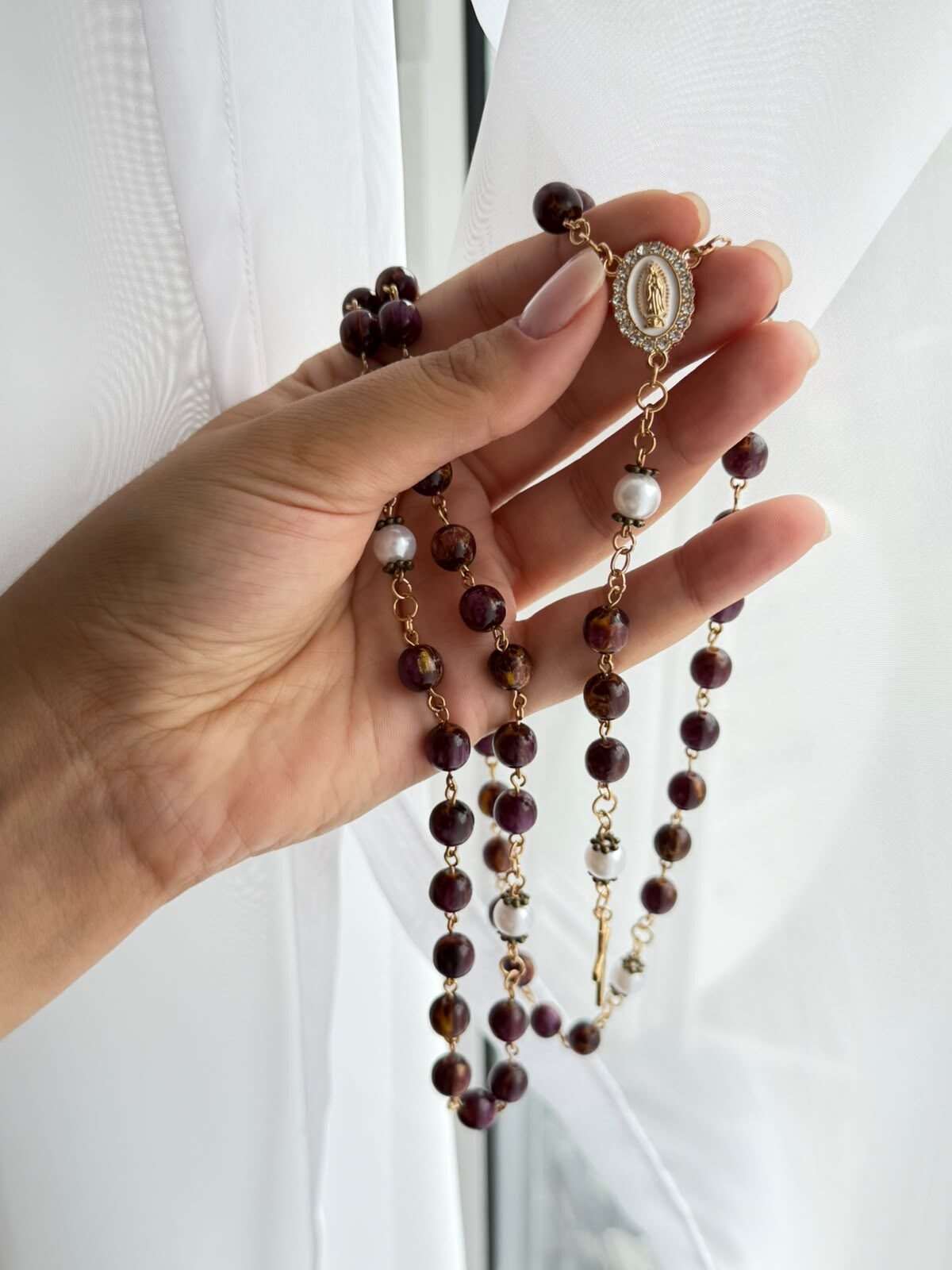 Deep plum beads streaked with warmer amber on warm-toned wire links, gathered across an open palm, a white round note at each turn and a small oval centre held between two fingers