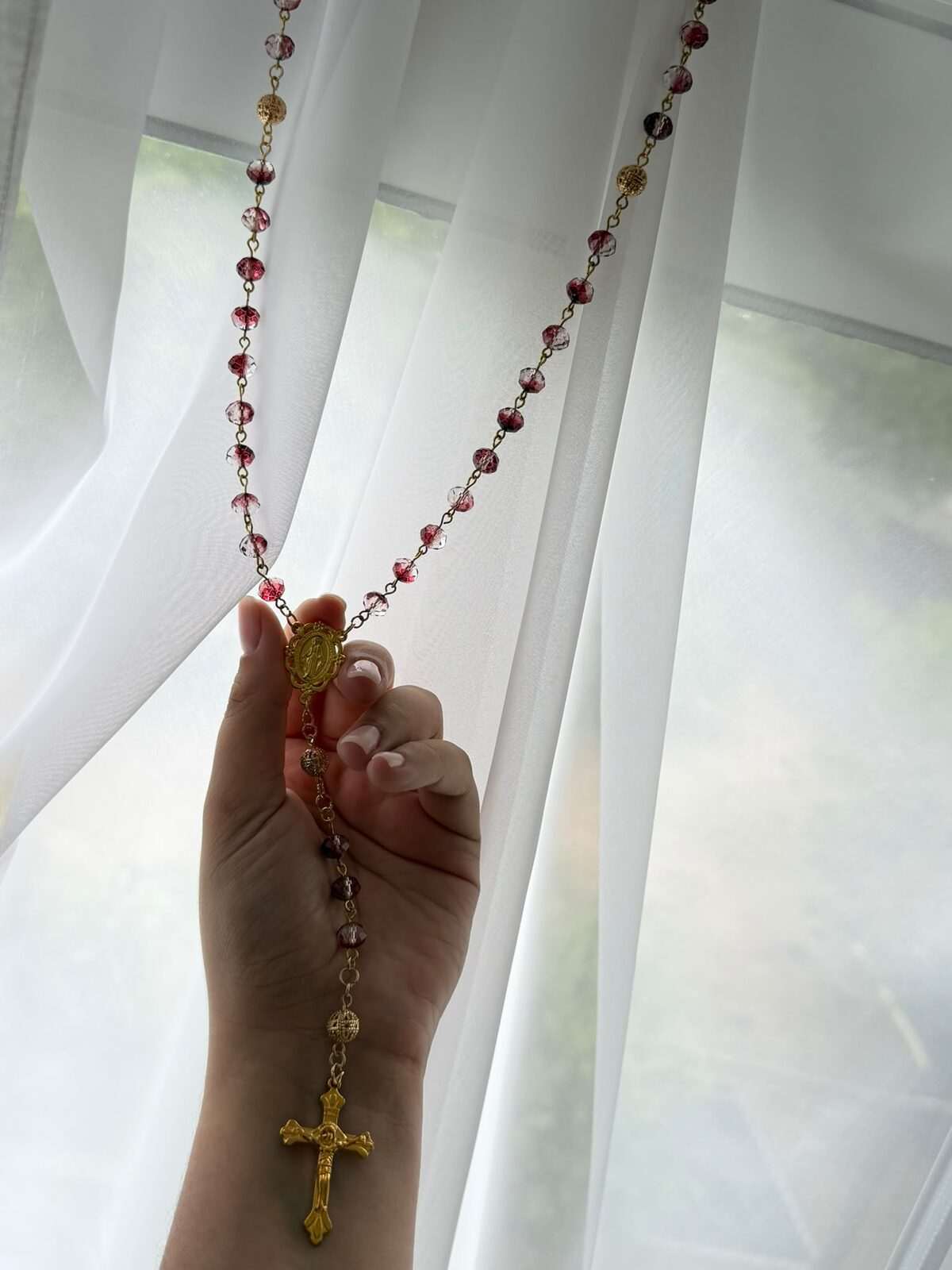 A long strand alternating deep wine and near-clear faceted beads on warm-toned wire links, hanging its full length from a raised hand, with worked round beads at the decades and a figured cross at the foot