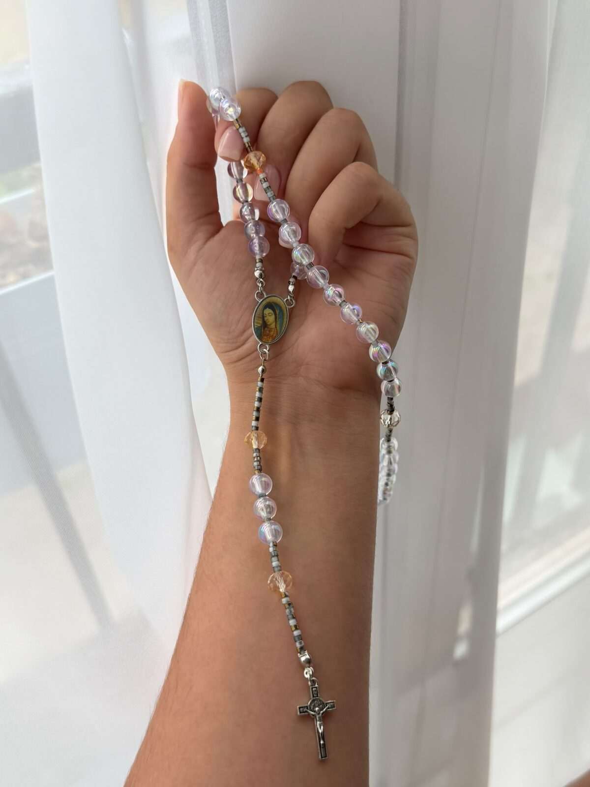 Pale violet iridescent round beads on fine pale-toned wire links falling across a closed hand and down a forearm, warm amber notes recurring, a small painted oval centre and a dark cross at the foot