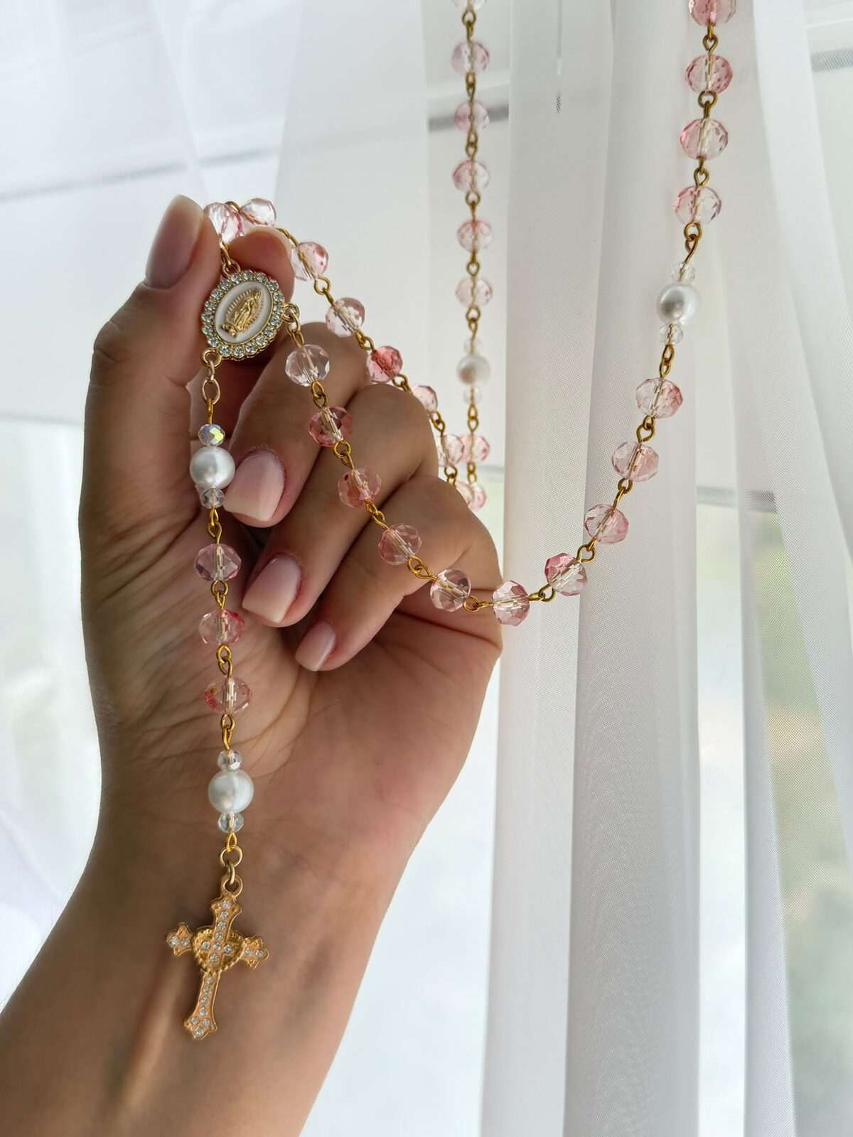 Pale pink and near-clear faceted beads alternating on warm-toned wire links, held at a small oval centre and falling in a wide loop, with white round notes at the decades and a worked cross at the foot