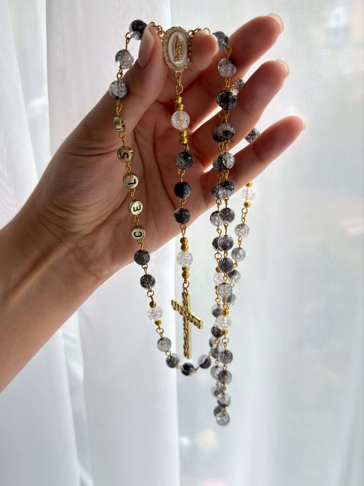 Dark speckled and near-clear beads alternating on warm-toned wire links across an open palm, a short line of small lettered beads reading downward, and a hammered cross hanging at the centre of the frame